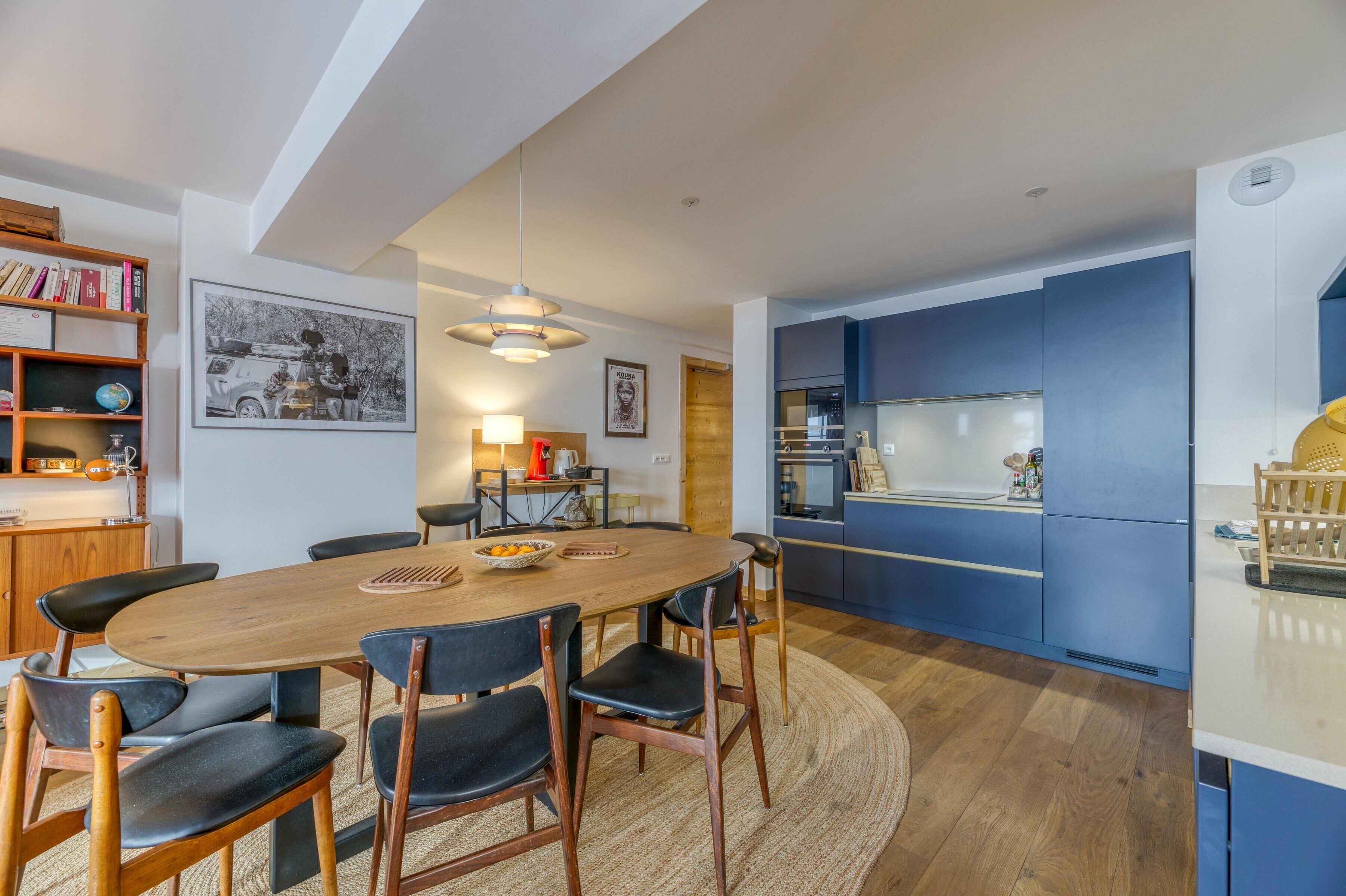 A modern kitchen with blue cabinets, a wooden dining table with chairs, and a hanging light fixture.