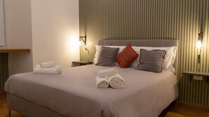 Una cama cuidadosamente tendida, con toallas enrolladas, un cabecero y dos lámparas de noche.