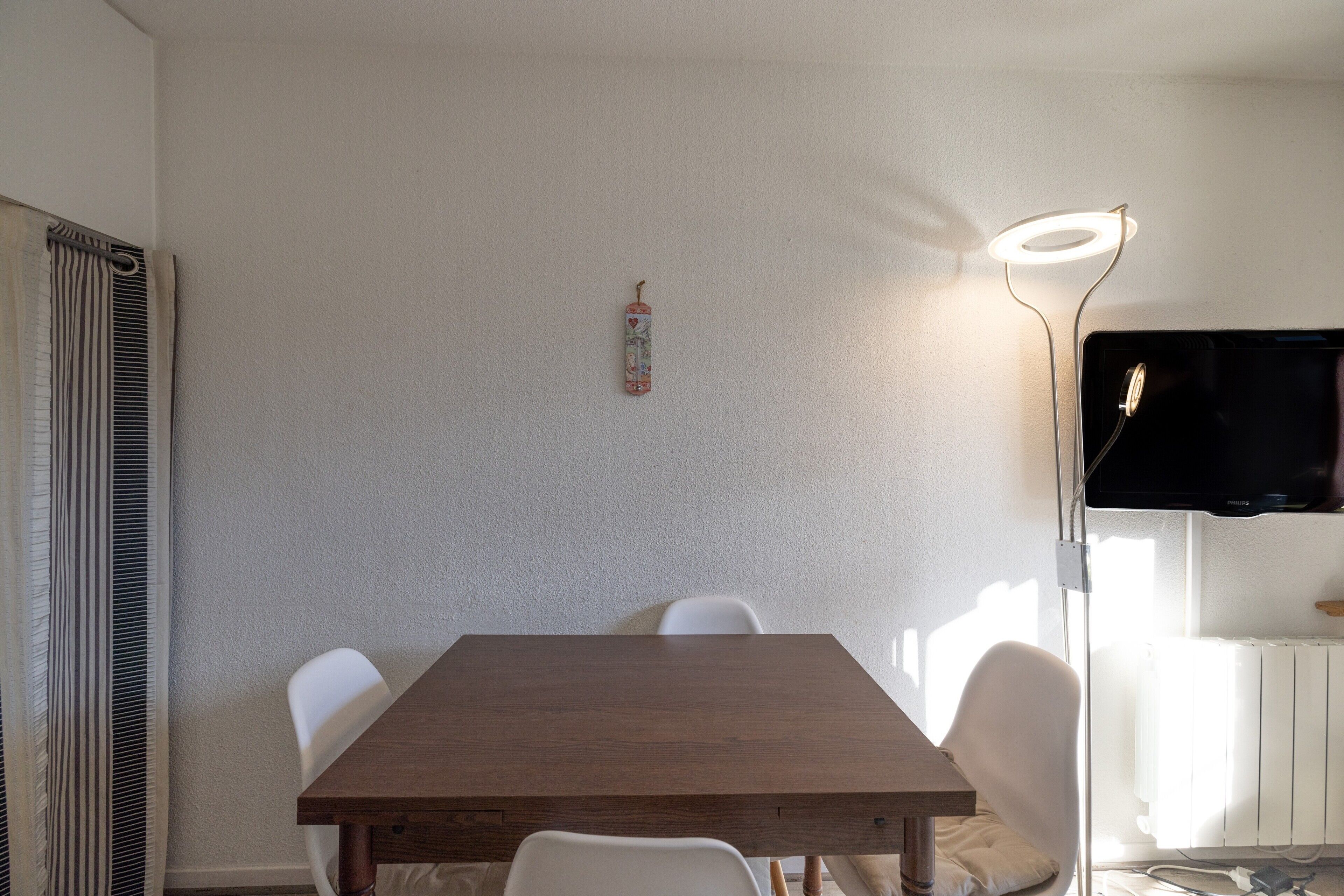 Un comedor con una mesa de madera, sillas blancas, un televisor montado en la pared y una lámpara de pie.