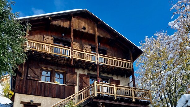 Een houten chalet met balkon, besneeuwde grond en een helderblauwe lucht.