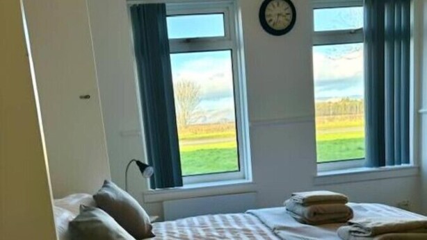 Une chambre à coucher avec un lit, deux fenêtres avec des rideaux, une horloge et une vue sur un paysage verdoyant.