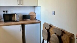 Une cuisine compacte avec des meubles blancs, un four à micro-ondes et un petit réfrigérateur. Des tabourets en bois sont fixés au mur, et un extincteur est accroché au mur.