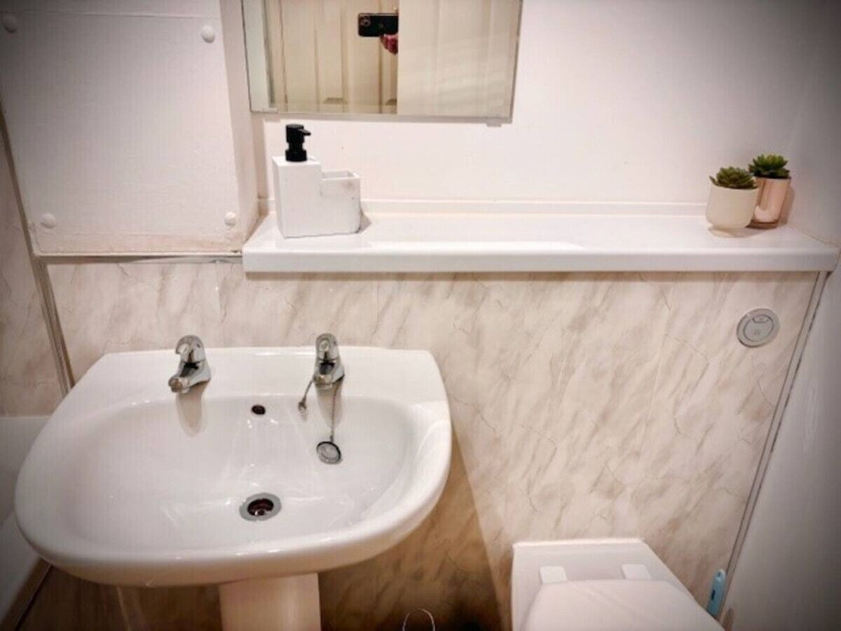 A bathroom with a white sink, two faucets, a mirror above the sink, a small shelf with a soap dispenser, and a toilet.