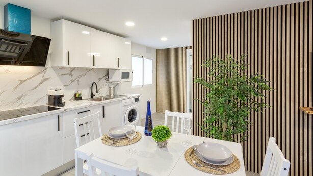 A modern kitchen with white cabinets, a built-in oven, and a microwave. There is a dining table set with plates, glasses, and a small plant. - Vacation Home 'Magma' with Private Terrace, Private Garden and Wi-Fi (Santa Brígida)