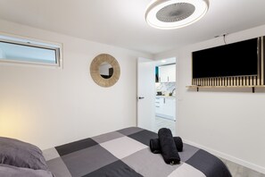 A modern bedroom with a checkered bedspread, a round mirror, a ceiling fan, a TV mounted on the wall, and a view into the adjacent kitchen.