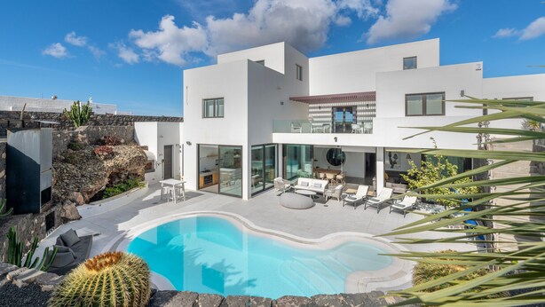 Una casa moderna de dos pisos con piscina, mobiliario exterior y vista al mar.