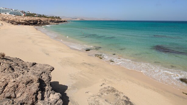A beach with clear turquoise water, sandy shore, rocky outcrops, and buildings in the background. - Vacation 'Home Sweet Home with Ocean View' with Sea View, Shared Pool and Wi-Fi (Costa Calma)
