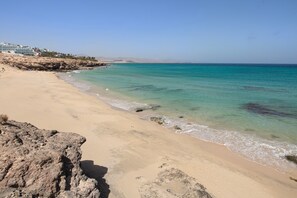 A beach with clear turquoise water, sandy shore, rocky outcrops, and buildings in the background.