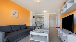 A living room with a sofa, coffee table, TV, and kitchen in the background. - Vacation 'Home Sweet Home with Ocean View' with Sea View, Shared Pool and Wi-Fi (Costa Calma)