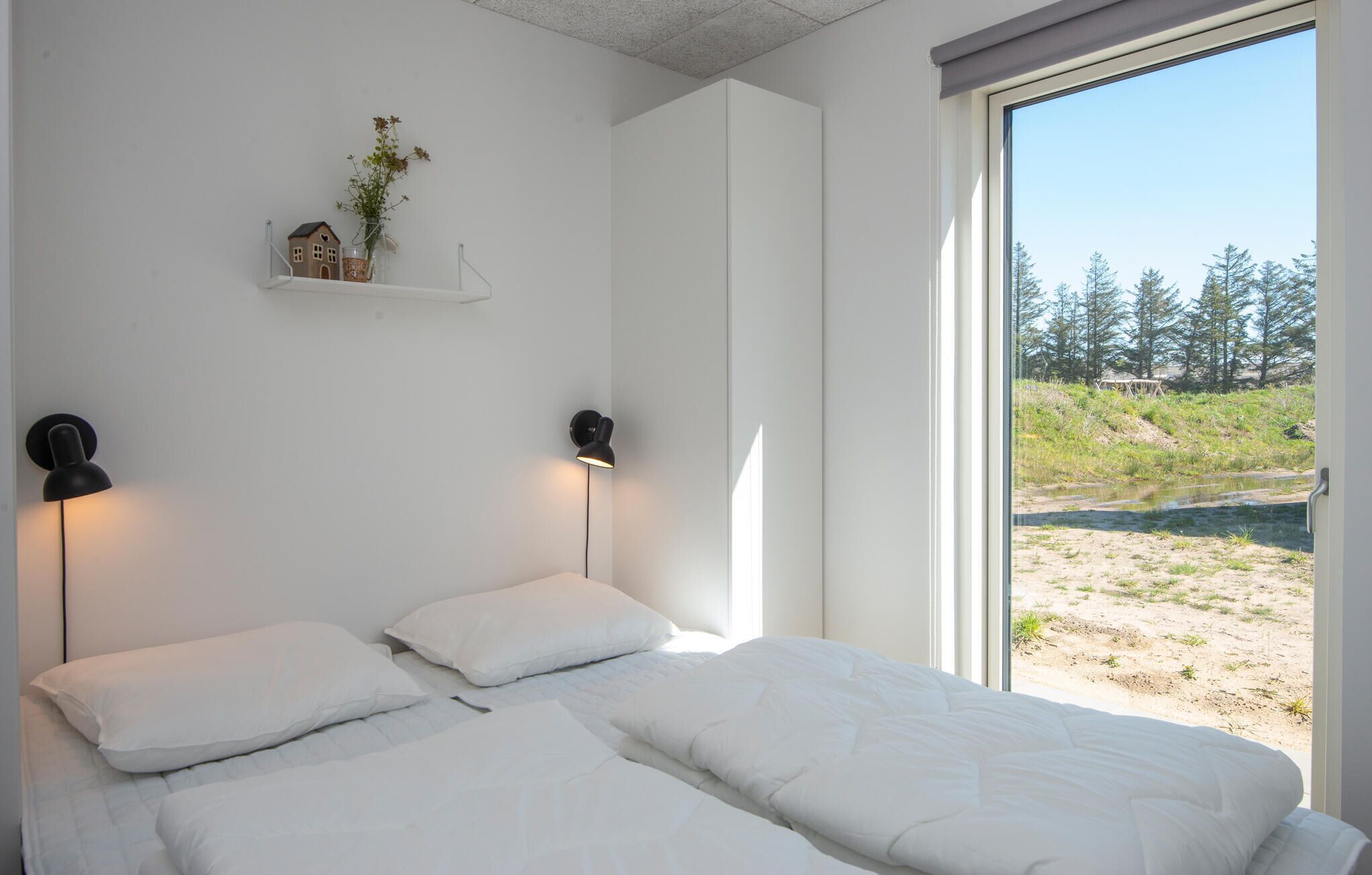 A bedroom with two beds, a small shelf with a house model and flowers, two wall-mounted lamps, and a large window with a view of a beach and trees.