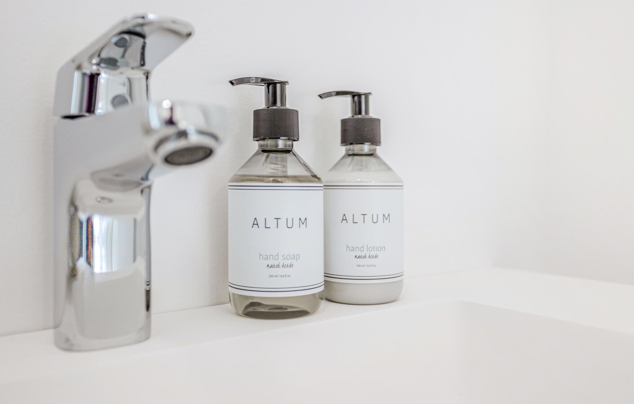 Two bottles of Altum hand soap and hand lotion, and a chrome faucet on a white surface.