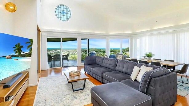 Ein modernes Wohnzimmer mit einem großen, grauen Ecksofa, einem Couchtisch und einem Flachbildfernseher. Ein Glastür öffnet sich zu einem Balkon mit Blick auf den Ozean und Palmen.