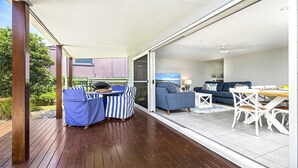Un espacio habitable de concepto abierto con un ventanal corredero que da a un patio. Se observa un sofá azul, una mesa de comedor blanca con sillas y una pequeña cocina visible a través del ventanal.