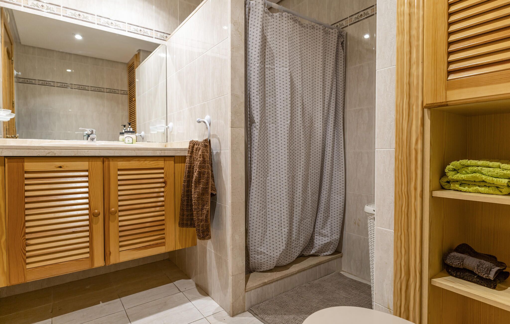 A bathroom with a wooden vanity, a shower with a patterned curtain, and a shelf with folded towels.