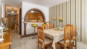 Un comedor con una mesa preparada para cuatro personas, rodeada de sillas de madera. Un arco de ladrillo conduce a una zona de cocina con armarios de madera y un microondas.