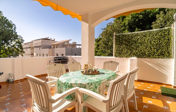 A patio with a table and chairs, a grill, and a view of buildings and greenery.