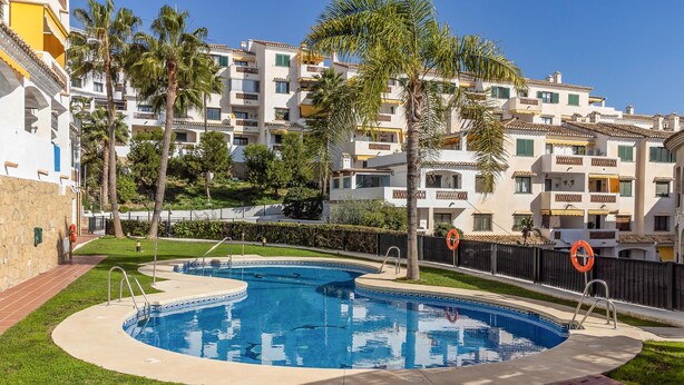 Una piscina rodeada por una valla, con palmeras y un complejo de edificios al fondo.