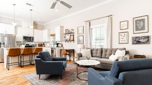 A modern living room with a sofa, armchairs, and a coffee table. There is a kitchen in the background with white cabinets and a dining area with a table and chairs.
