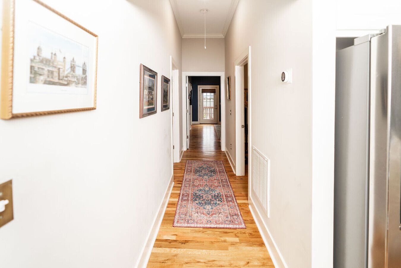 A hallway with wooden flooring, a patterned rug, framed pictures on the walls, and a refrigerator on the right.