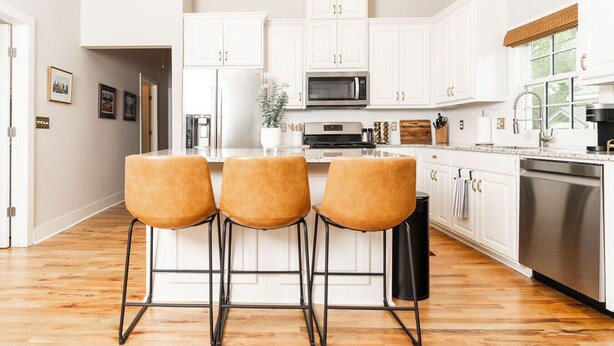 A modern kitchen with white cabinets, a central island with stools, stainless steel appliances, and a wooden floor.
