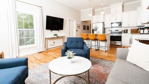 A modern living room with a blue sofa, a grey sofa, and a round coffee table with a plant. There is a TV mounted on the wall, a kitchen area with white cabinets and a microwave, and a patterned rug on the floor.