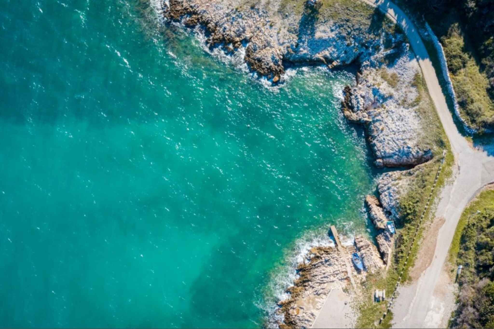 Vue aérienne d’une zone côtière avec des eaux turquoise limpides, un terrain rocheux et un sentier pavé longeant la côte.