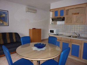 Um espaço compacto com mesa de vidro redonda, cadeiras azuis, sofá, cozinha compacta com micro-ondas e armário.