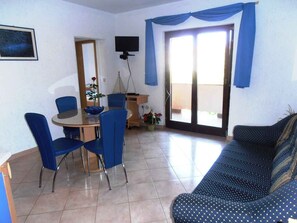Uma sala de estar com sofá, mesa de jantar e cadeiras, TV fixada na parede e porta de vidro corrediça para uma varanda.