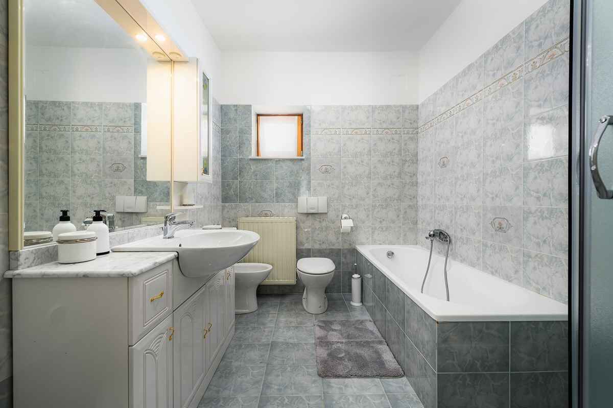 A modern bathroom with a sink, toilet, and bathtub, featuring marble tiles and a small window.
