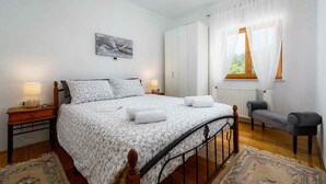 Un dormitorio con cama, mesitas de noche, una silla, un ventanal con cortinas y un radiador.