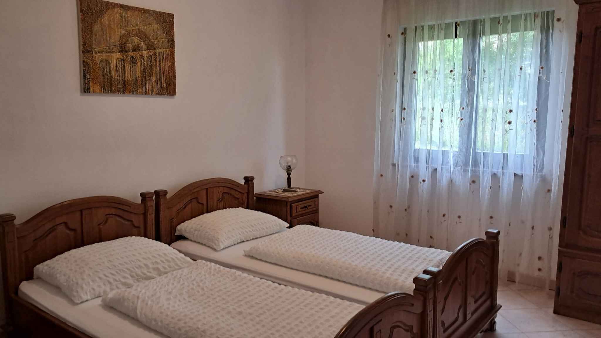 A bedroom with two wooden beds, a nightstand with a lamp, a painting on the wall, and a window with sheer curtains.