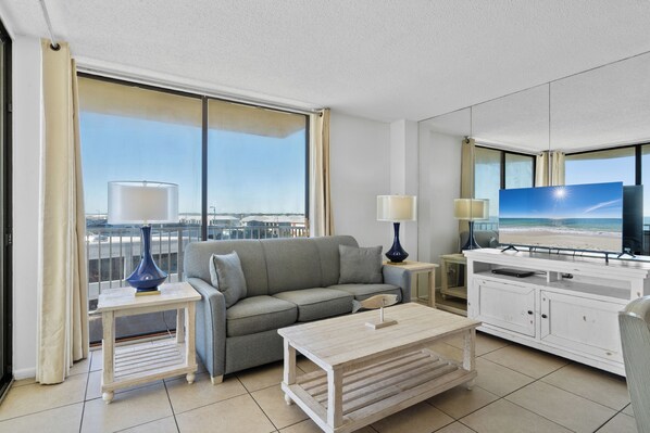 Una habitación de hotel moderna con sofá gris, una mesa de centro de madera, un televisor sobre un mueble blanco y un ventanal con vistas a la playa.