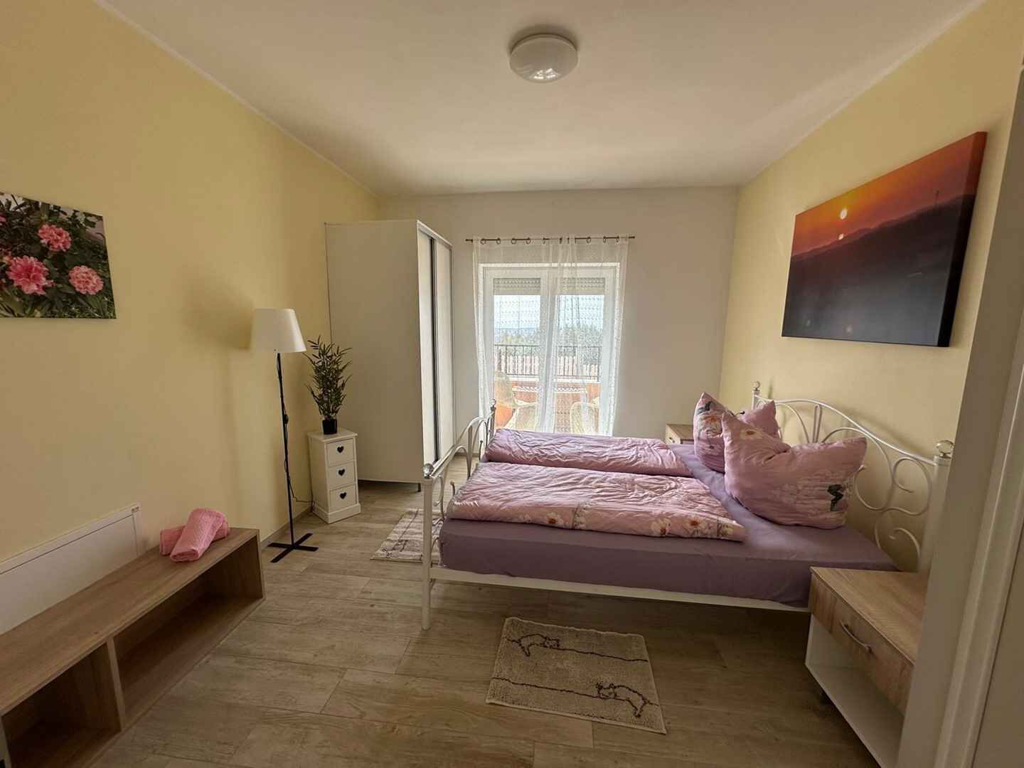 A bedroom with a bed, a bedside table, a lamp, a bench, and a window with a view.