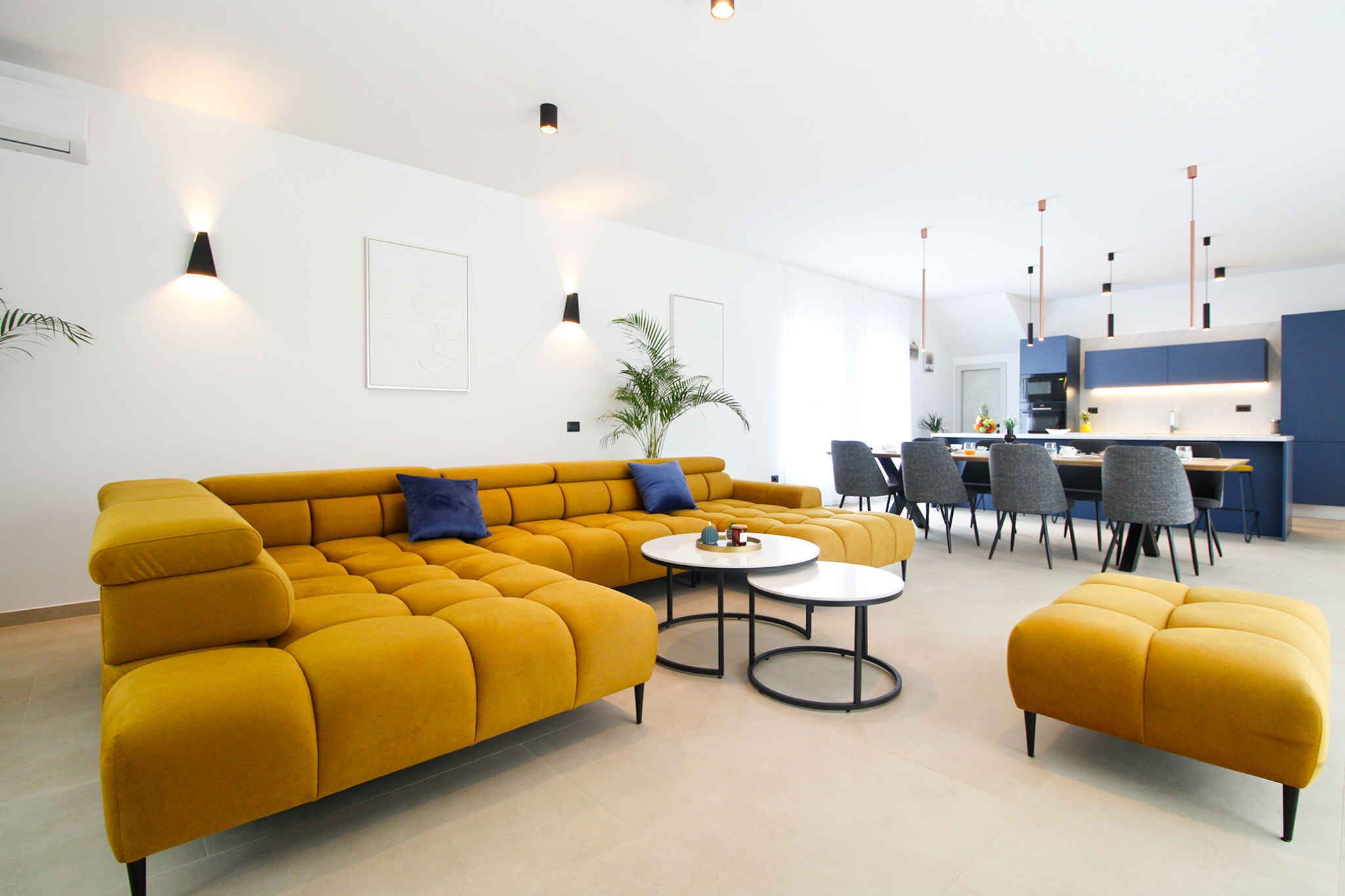 Een moderne woonkamer met een mosterdgele bank, een ronde salontafel en een bijpassend voetenbankje. Op de achtergrond een eethoek met een tafel en stoelen.