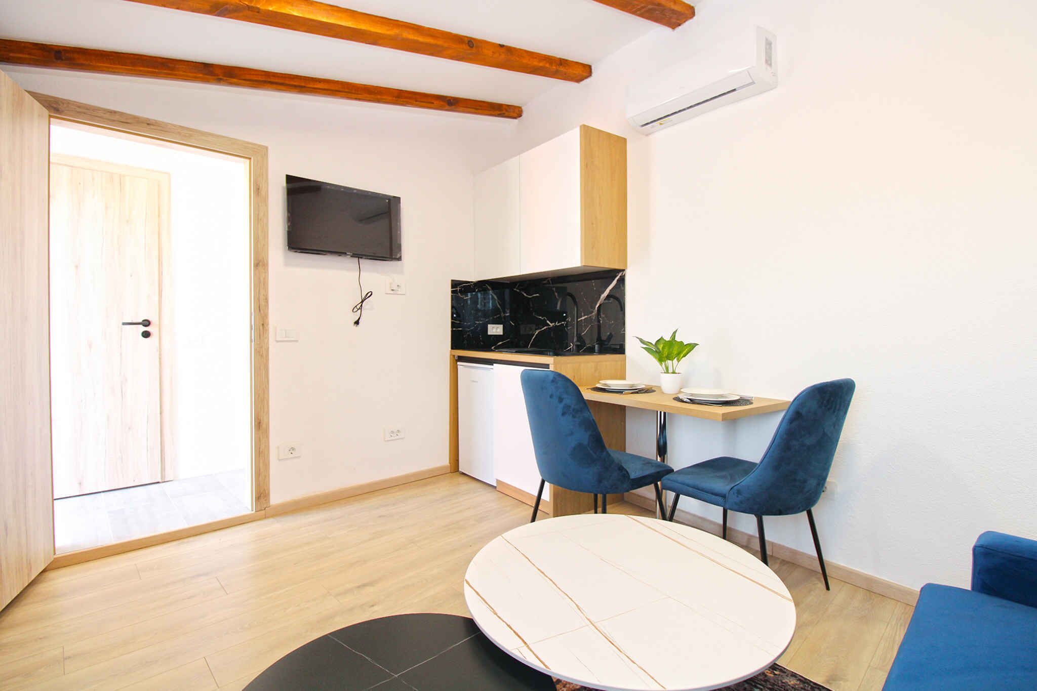 Uma cozinha moderna com bancada preta, duas cadeiras azuis, uma mesa pequena e uma TV fixada na parede.