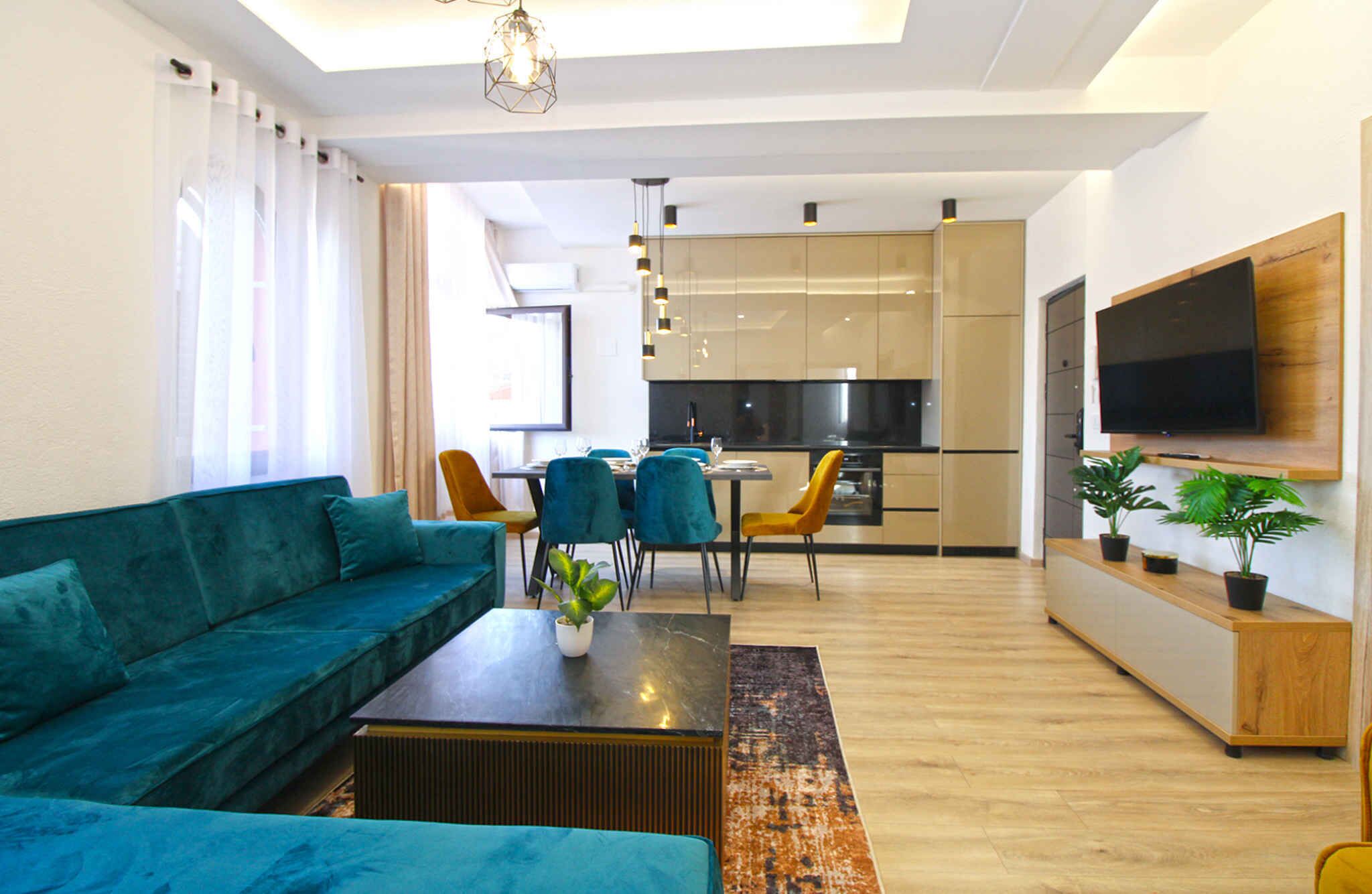 Ein modernes Wohnzimmer mit einem türkisfarbenen Sofa, einem Couchtisch und einem Essbereich mit Stühlen. Es gibt eine Küche mit Fernseher und einem großen Fenster.
