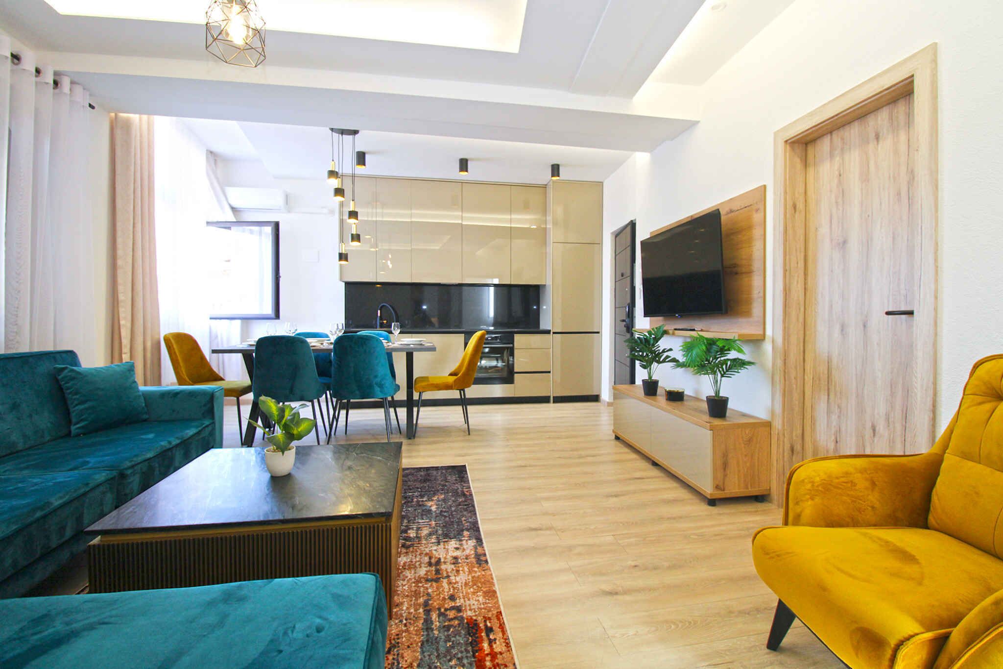 A modern living room with a sofa, armchair, and coffee table. There is a dining area with chairs and a table. A kitchen with cabinets and a TV is visible in the background.