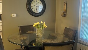 Zona pranzo con un tavolo rotondo in vetro, quattro sedie, un vaso con fiori gialli, una sveglia a parete e un elemento decorativo in metallo.