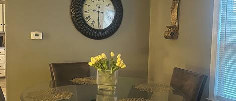 Un comedor con una mesa redonda de vidrio, cuatro sillas, un florero con flores amarillas, un reloj de pared y un adorno metálico.