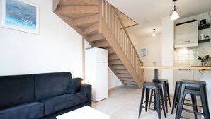 A modern living room with a black sofa, a white coffee table, and a wooden staircase leading to a kitchen area. - Beautiful duplex villa for 5 pers, air-conditioned (Sérignan)