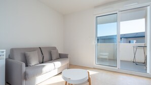 Un salon minimaliste avec un canapé gris, une petite table ronde et une porte-fenêtre coulissante donnant sur un balcon extérieur.