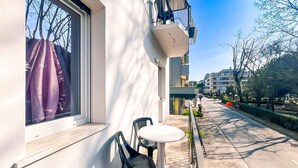 A balcony with a table and chairs, a window with a curtain, and a clear view of the street and buildings in the background.