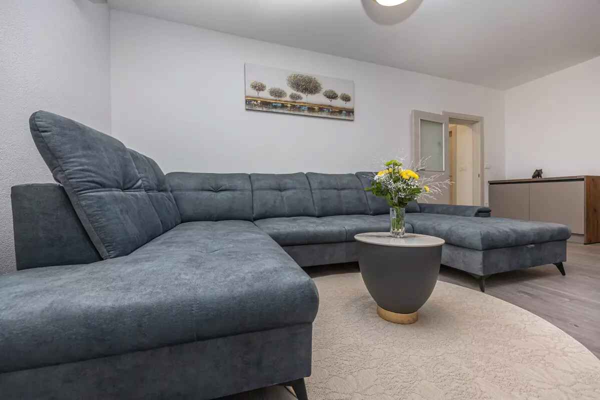 Ein modernes Wohnzimmer mit einem grauen Ecksofa, einem runden Couchtisch mit Blumenvase und einem Bild an der Wand.