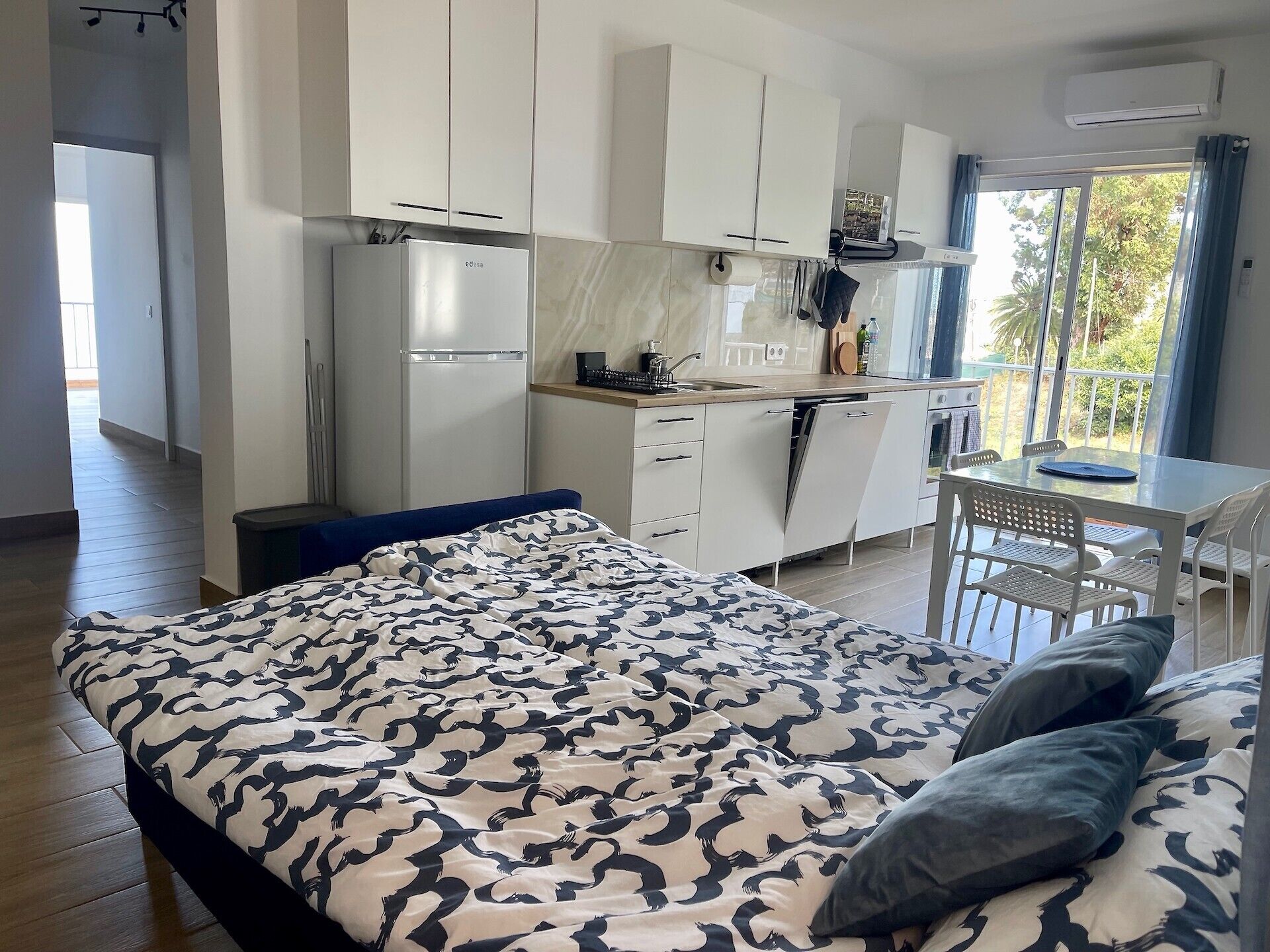 Un dormitorio moderno con una colcha estampada, una pequeña kitchenette y un balcón con mobiliario exterior.