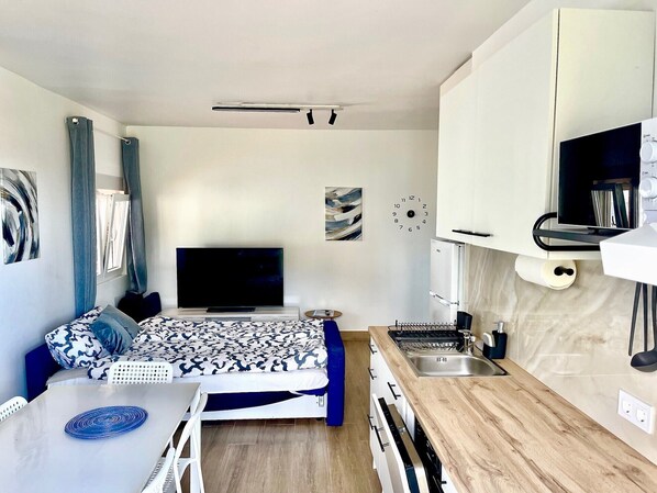 Un espacio habitable compacto con cama, televisor de pantalla plana, zona de cocina con lavaplatos y estufa, y un reloj en la pared.