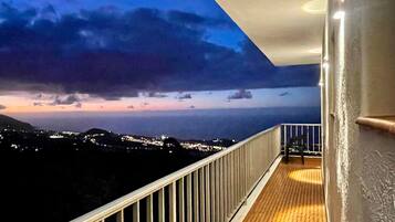 Un balcón con barandilla, piso de madera y vista a un paisaje urbano al anochecer.