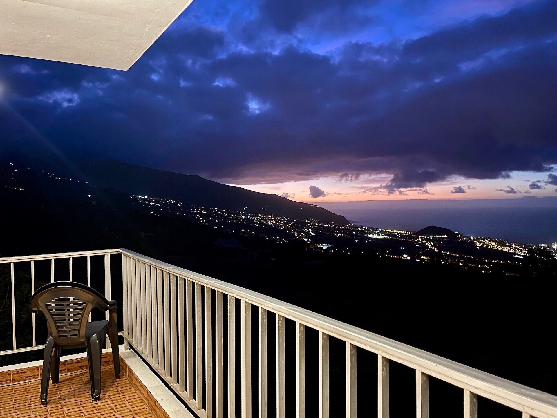 Ein Balkon mit Blick auf eine Stadt im Abendrot, ein Stuhl und ein Geländer.