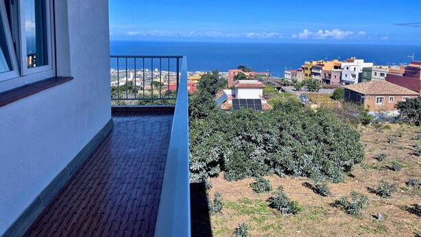 A balcony with a view of the sea, a tiled floor, and a white building.
