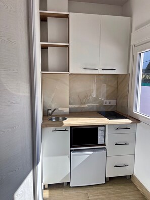 Una cucina compatta con mobili bianchi, forno a microonde integrato, lavello e finestra.