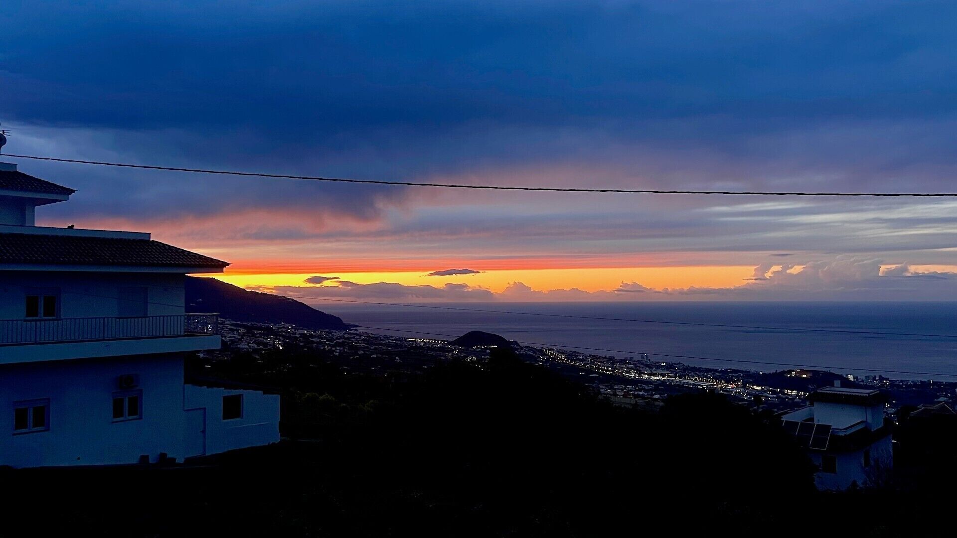 A coastal cityscape at sunset with a prominent building on the left and a view of the ocean.